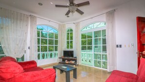 A spacious living room with a red sofa, a coffee table, and a TV. Large windows with white curtains provide ample natural light and views of greenery outside. - Apartment Castello 1B (Playa del Carmen)