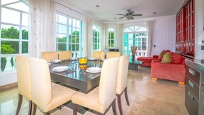 A spacious dining area with a large table, chairs, and a bowl of lemons. There is a red sofa and a kitchen area with cabinets and appliances in the background. - Apartment Castello 1C (Playa del Carmen)