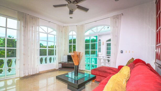 A spacious living room with a red sofa, a glass coffee table, and large windows offering a view of greenery. - Apartment Castello 1C (Playa del Carmen)