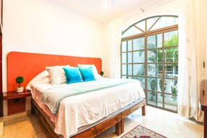A bedroom with a large bed, a wooden headboard, a nightstand, a small potted plant, and a large window with a view of greenery.