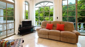Un salón con un sofá color crema, un televisor de pantalla plana sobre un mueble bajo y ventanas amplias con cortinas.