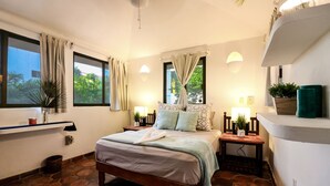 A bedroom with a bed, two bedside tables, a window with curtains, a wall-mounted shelf with plants, and a stone-tiled floor. - Villa 4 Soles (Playa del Carmen)