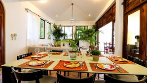 A dining area with a long table set for six, surrounded by chairs, with a sofa and plants in the background. - Villa 4 Soles (Playa del Carmen)