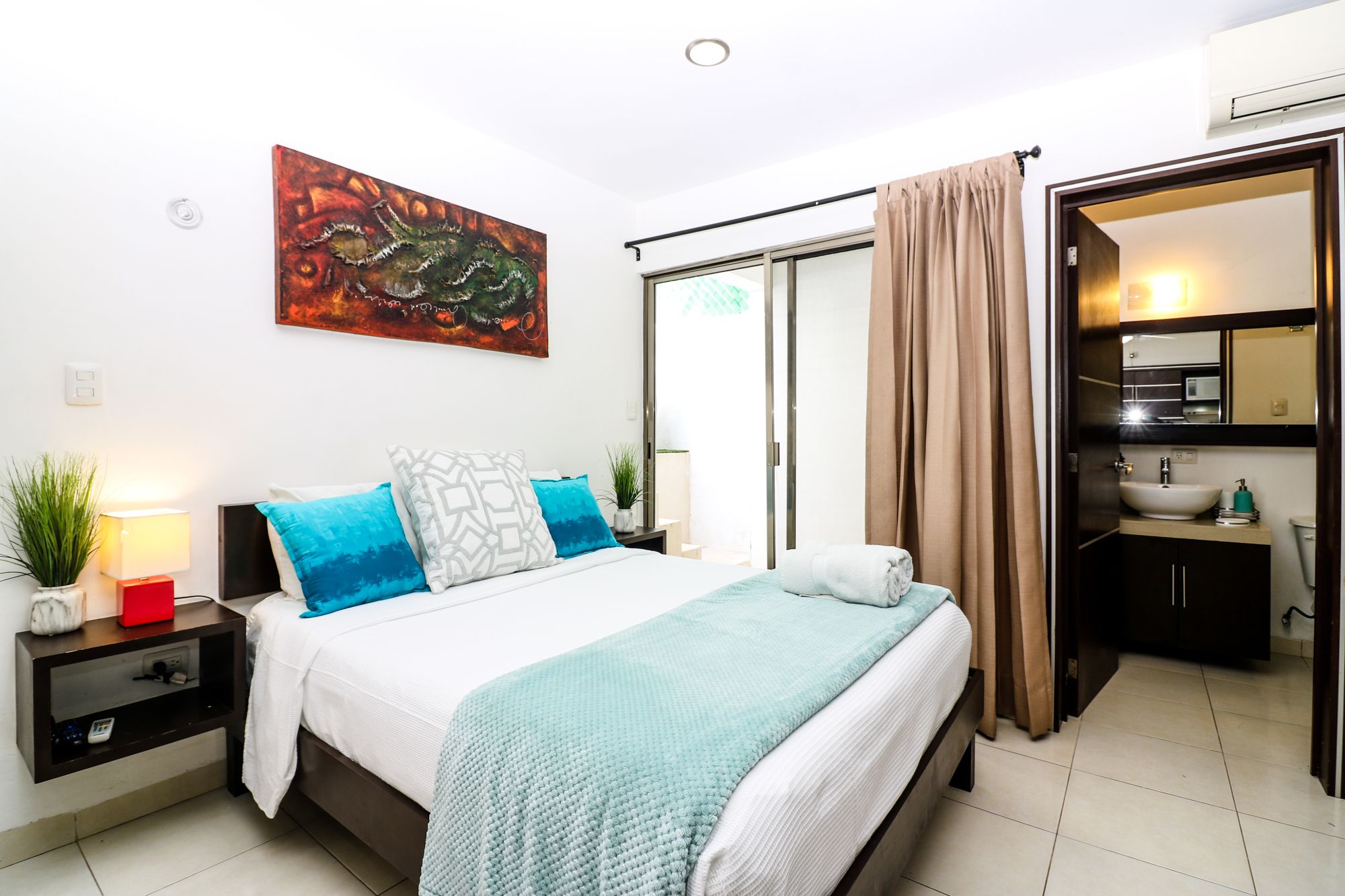 Une chambre d’hôtel moderne, avec un lit, des tables de chevet, un tableau et une salle de bain visible à travers une porte ouverte.