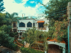 A two-story house with a balcony, surrounded by lush greenery and a clear sky.