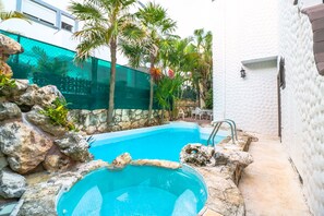 A swimming pool with a stone border, surrounded by rocks and palm trees, with a white building in the background.