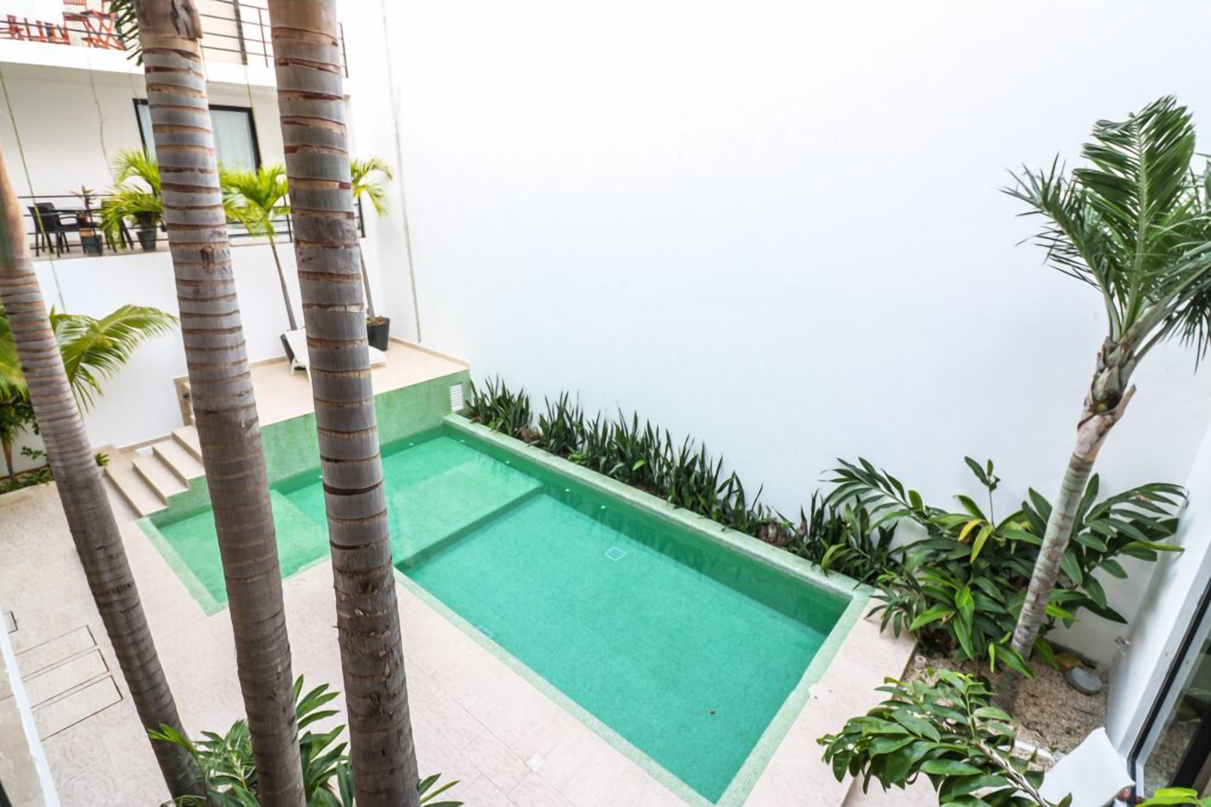 Ein rechteckiger Swimmingpool, umgeben von Palmen und Pflanzen, mit einem Balkon und einer Sitzgelegenheit im Hintergrund.
