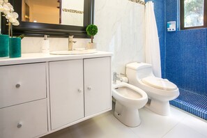 A modern bathroom with a white vanity, two toilets, a bidet, a small potted plant, and a blue-tiled shower area. - Casa 20 (Playa del Carmen)