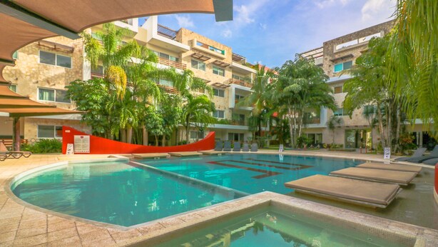 A swimming pool with lounge chairs and palm trees, surrounded by multi-story buildings. - Apartment Sabbia B401 (Playa del Carmen)