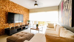 A modern living room with a brick wall, a flat-screen TV, a sofa, a coffee table, and a balcony with yellow chairs. - Apartment Sabbia B401 (Playa del Carmen)