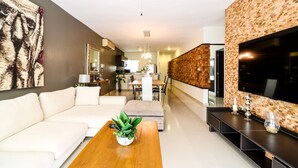 A modern living room with a sofa, coffee table, and a TV. There is a kitchen in the background. - Apartment Sabbia B401 (Playa del Carmen)