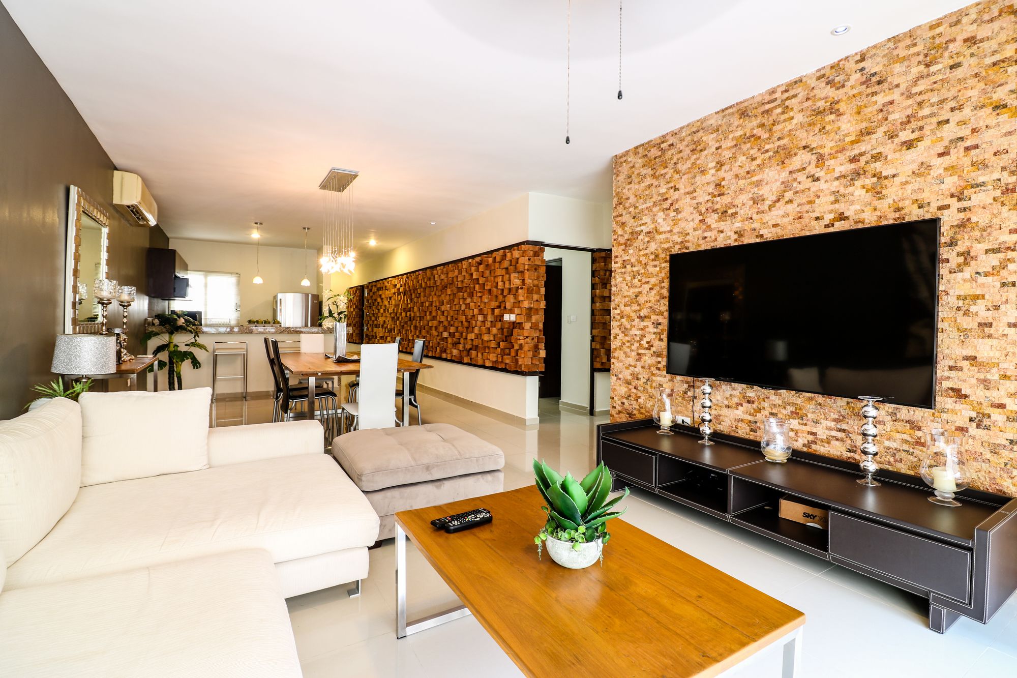 A modern living room with a sectional sofa, a wooden coffee table, a TV mounted on a textured wall, and a dining area in the background.