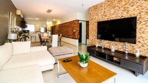 A modern living room with a sectional sofa, a wooden coffee table, a TV mounted on a textured wall, and a dining area in the background. - Apartment Sabbia B401 (Playa del Carmen)