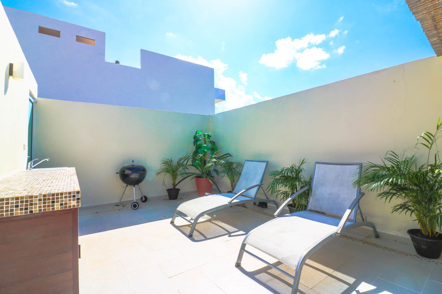 A patio with a tiled floor, a barbecue grill, several potted plants, and two lounge chairs.