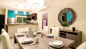 A dining area with a wooden table set for four, surrounded by white chairs. A large mirror with a decorative frame hangs on the wall, and a chandelier is visible above the table. - Apartment Sabbia B402 (Playa del Carmen)