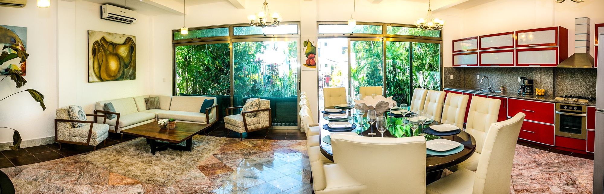 A modern living room with a sofa, armchairs, a coffee table, and a dining area with a table set for four. There is a kitchen with red cabinets and a stove. Large windows provide a view of greenery outside.