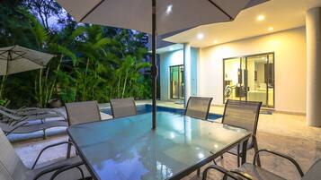 An outdoor pool area with a glass table and chairs, surrounded by palm trees and a modern house in the background.