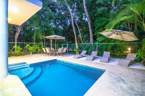 Une piscine entourée d’une clôture et d’une végétation luxuriante, avec des chaises longues et des parasols à proximité.