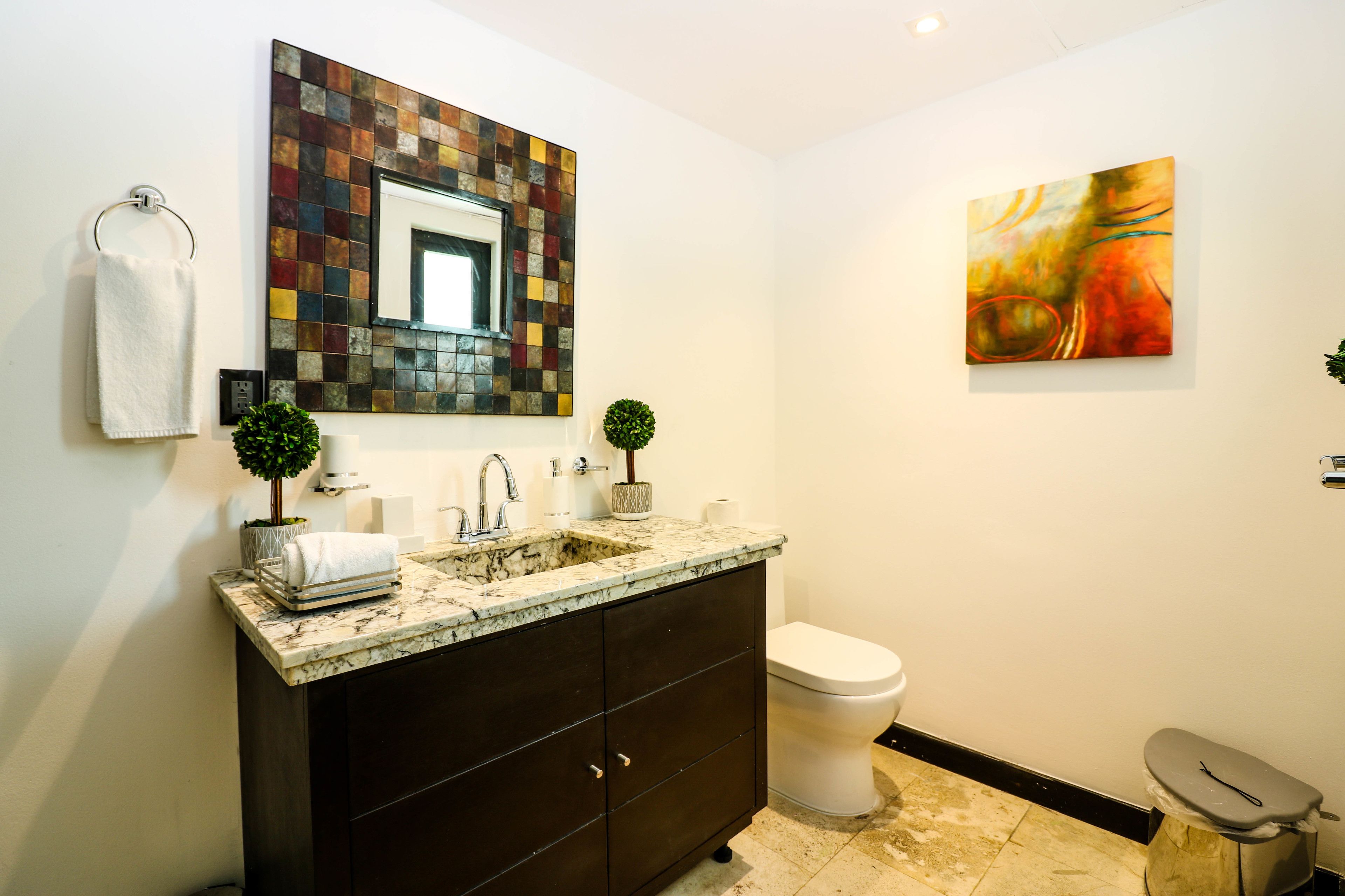 Une salle de bain moderne avec un lavabo en marbre, des meubles sombres, des toilettes blanches, une petite plante en pot et une peinture abstraite aux couleurs vives accrochée au mur.
