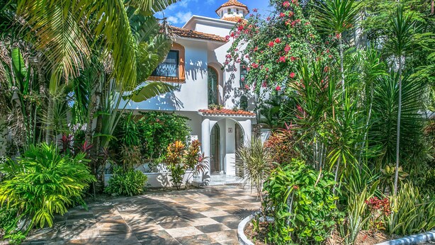 A white building with a tiled roof, surrounded by lush tropical plants and a checkered pathway leading to the entrance. - Villa Compound (Playa del Carmen)
