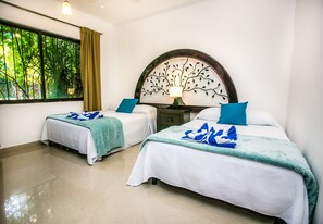 Una habitación de hotel con dos camas, una mesita de noche con lámpara, un ventanal con cortinas y un cabecero decorado con motivos de árboles.