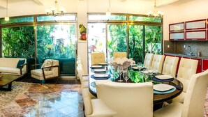 A modern living room with a sofa, armchairs, a coffee table, and a dining area with a table set for four. There is a kitchen with red cabinets and a stove. Large windows provide a view of greenery outside. - Villa Compound (Playa del Carmen)