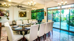 A dining area with a round table, white chairs, and a view of a garden through large windows. - Villa Compound (Playa del Carmen)