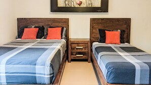 Une chambre avec deux lits, chacun doté d’une tête de lit en bois et d’une couverture à carreaux. Une petite table de chevet en bois, avec des tiroirs, se trouve entre les deux lits. Au-dessus des lits, un tableau encadré est accroché au mur.