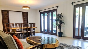 Un salon avec une table basse en bois, un canapé recouvert d’un plaid, une table à manger avec des chaises et une grande plante en pot.