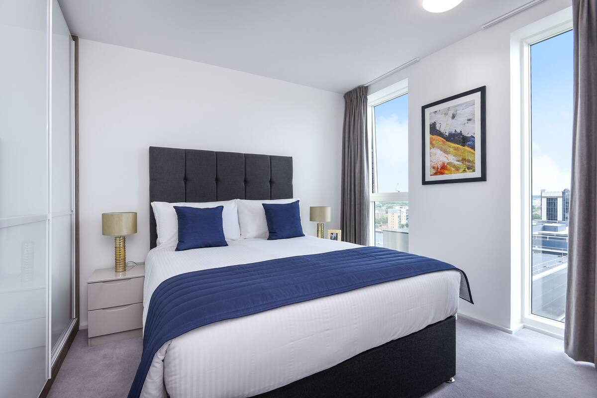 Quarto de hotel moderno com uma cama grande, mesa de cabeceira com abajur, janela com vista para a cidade e um quadro na parede.