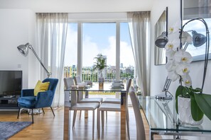 A modern living room with a glass dining table, blue and yellow chairs, a TV, and a large window with a city view. - Millharbour Residences (London)
