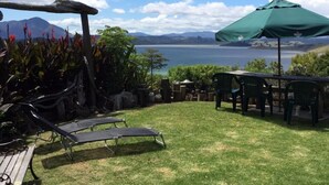 A patio with a view of a lake and mountains, featuring lounge chairs, a table, and an umbrella. - MUDBRICK BARN MUDENZA VU (Opononi)