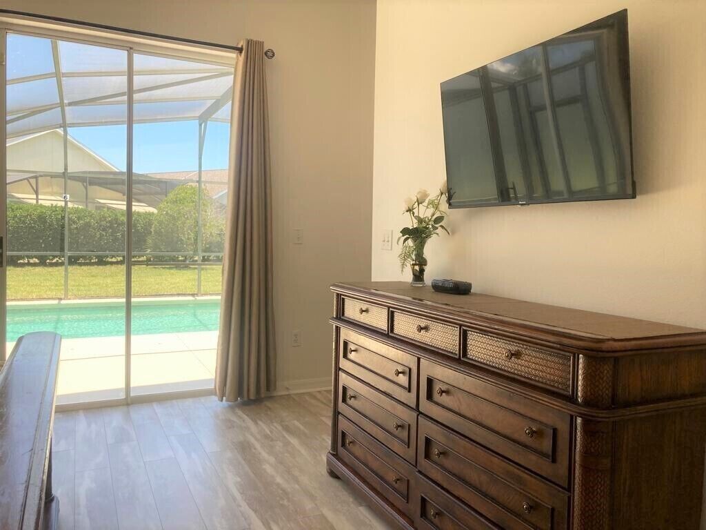 A room with a large sliding glass door leading to a pool, a wooden dresser with drawers, and a flat-screen TV mounted on the wall.