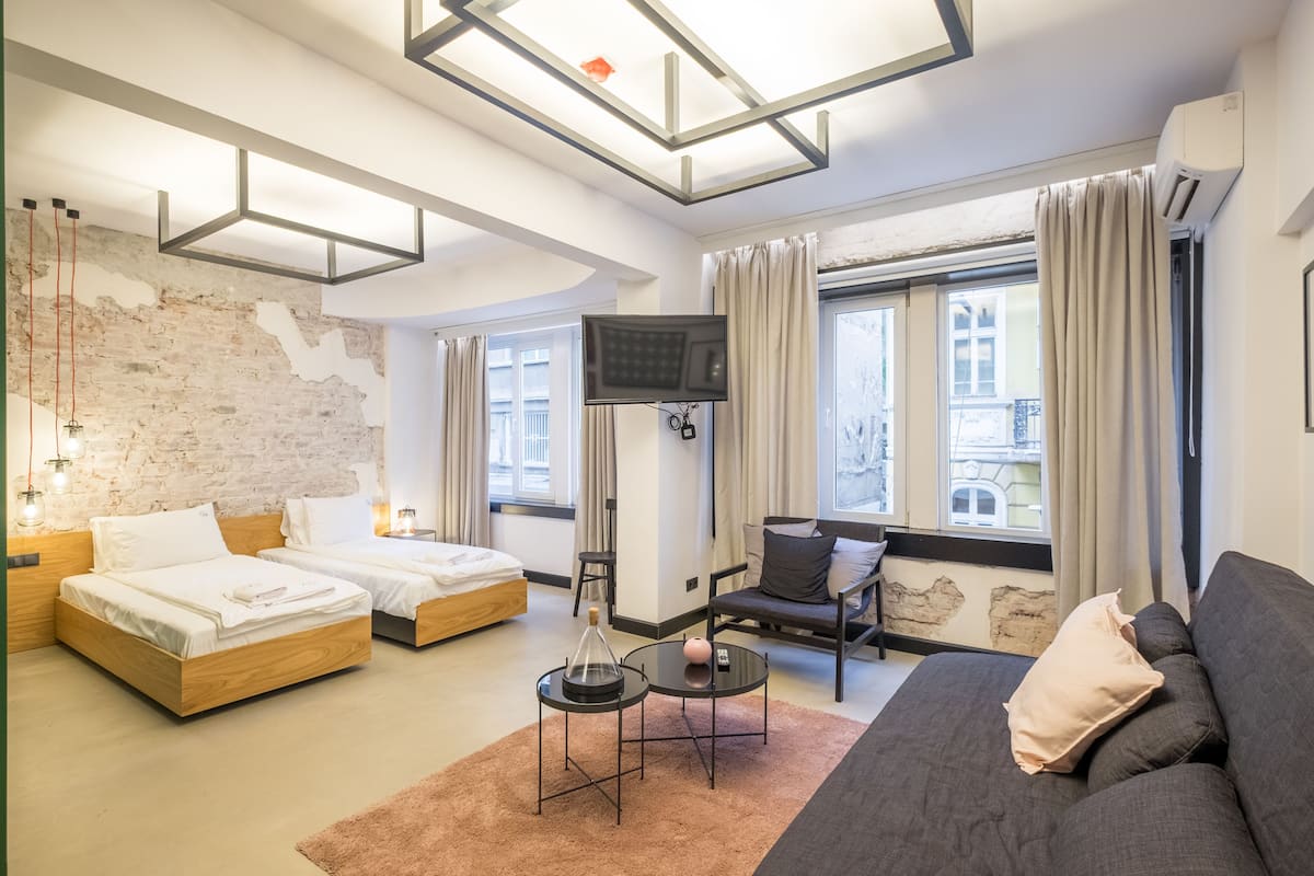 Habitació d'hotel moderna amb dos llits de fusta, un sofà gris, una televisió i una finestra gran amb cortines.