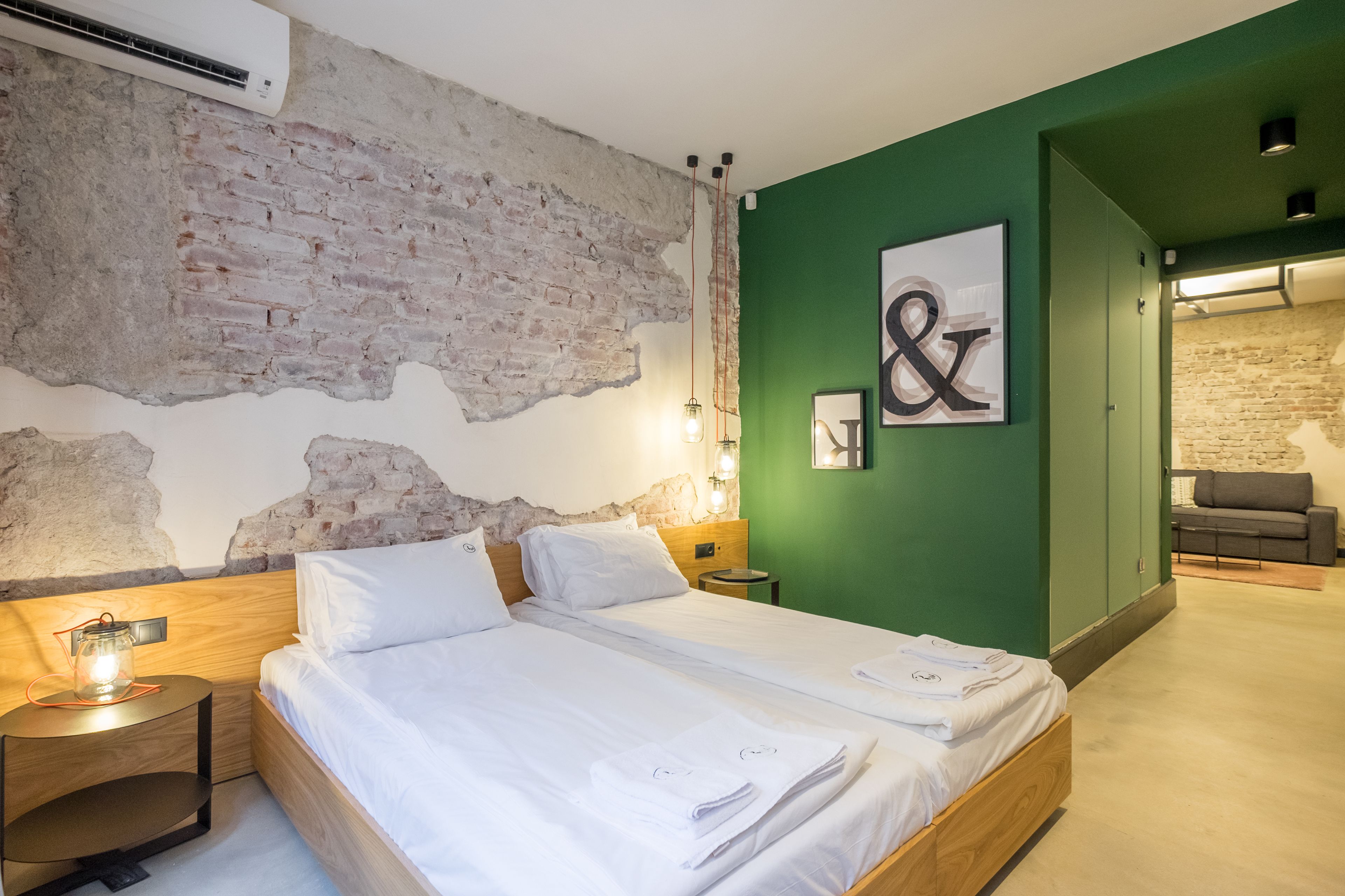 Ein modernes Hotelzimmer mit Backsteinwand, grünen Wänden, einem hölzernen Bett, einem Nachttisch mit Lampe und einem Sofa im angrenzenden Raum.