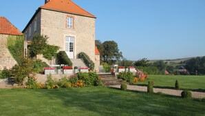 A stone building with a red-tiled roof, a staircase leading to the entrance, and a well-maintained garden with flowers and shrubs.