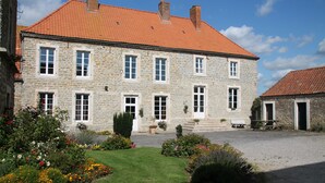 A stone building with a red-tiled roof, multiple windows, and a gravel courtyard with flower beds.
