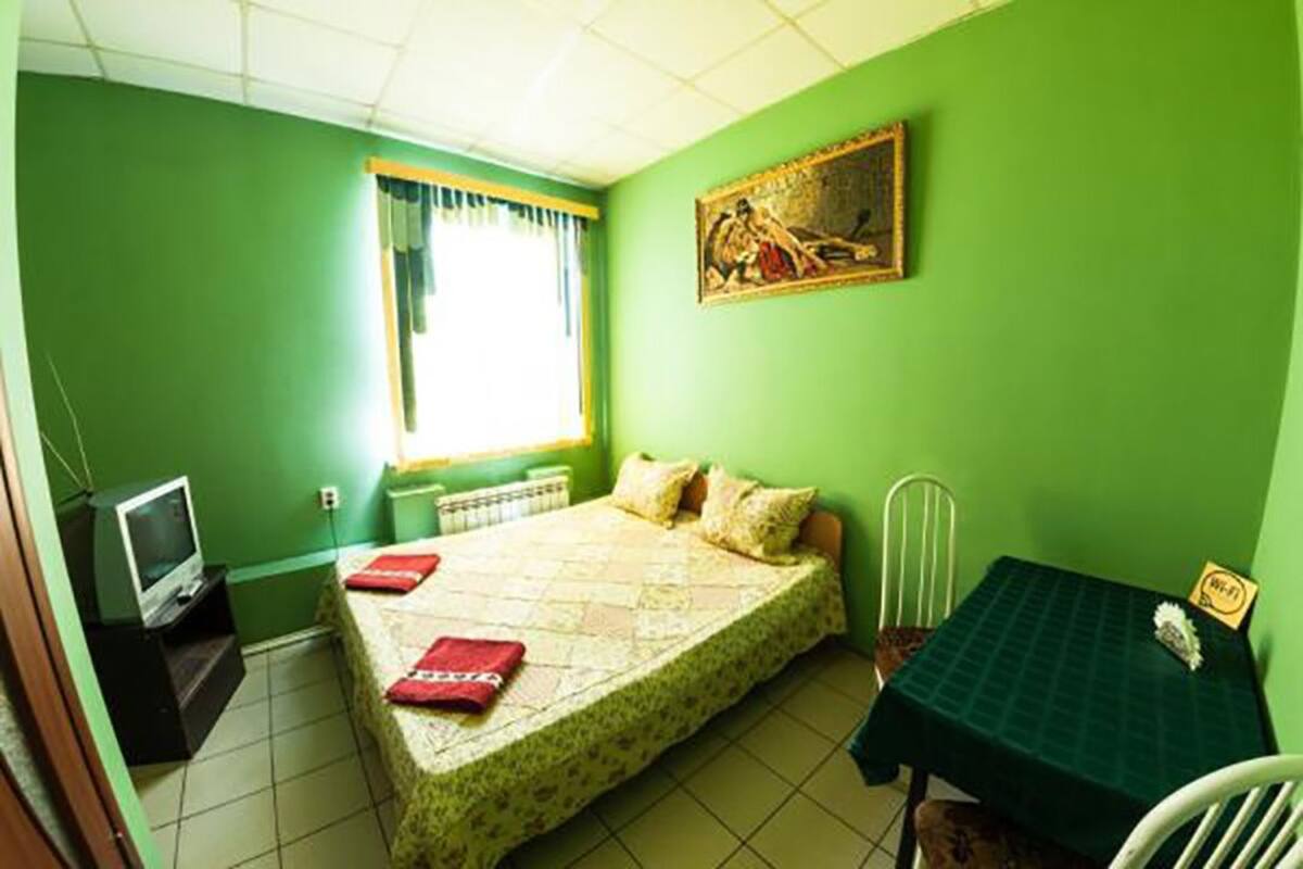 Una habitación con cama, una mesita pequeña con mantel verde, una silla, un televisor y una ventana con cortinas.