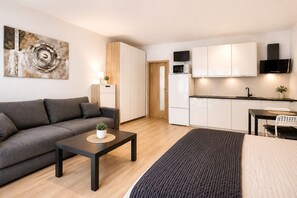 Un appartement moderne avec un canapé gris, une petite table sur laquelle est posée une plante, un coin cuisine doté de meubles blancs et un grand tableau abstrait accroché au mur.