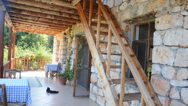 A stone building with a wooden staircase leading to a balcony. There are tables and chairs outside, and a cat is lying on the floor. - Telemen'in Evi Pansiyon (Demre)