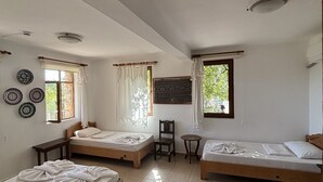 A room with three wooden beds, a small table, and a chair. There are windows with curtains and a view of greenery outside. - Telemen'in Evi Pansiyon (Demre)