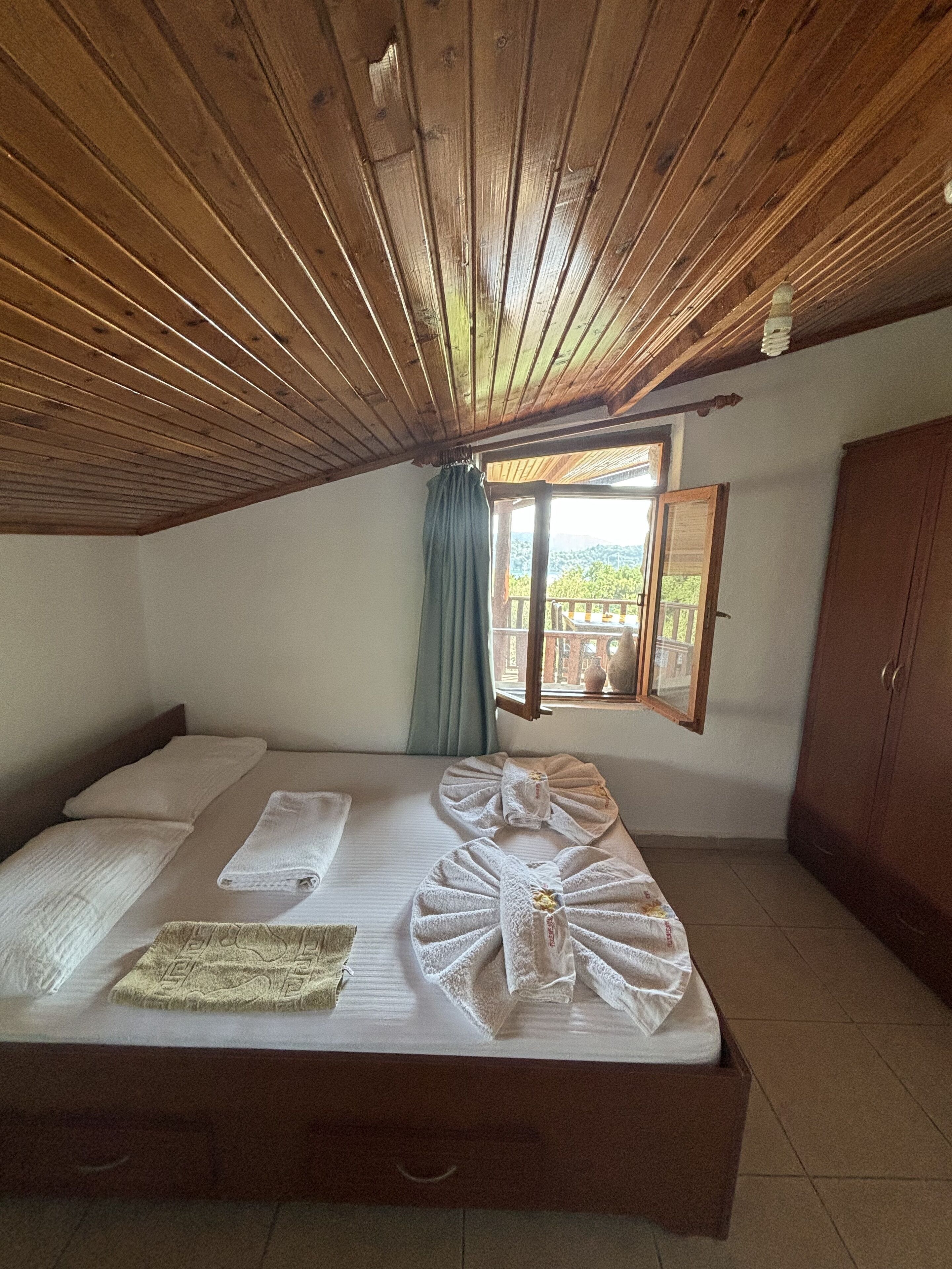 A bedroom with a wooden ceiling, a bed with neatly folded towels, a window with a view, and a wardrobe.