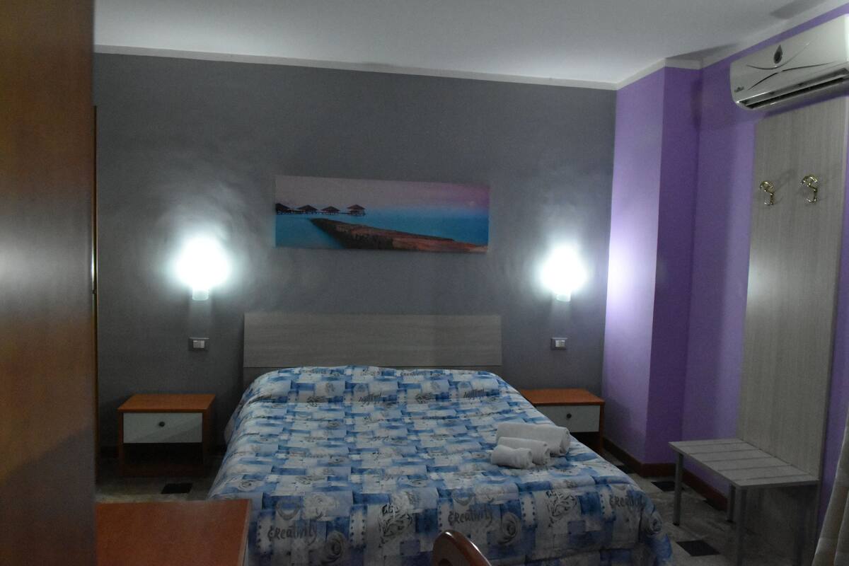 A bedroom with a bed, two bedside tables, a nightstand, a wall-mounted air conditioner, and a painting of a coastal scene.