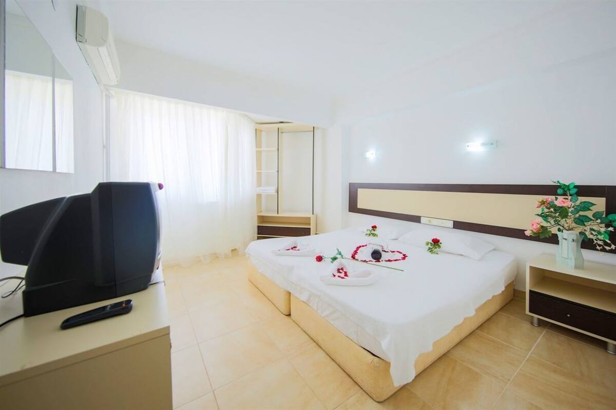 Quarto de hotel com cama, televisão, escrivaninha e mesa de cabeceira.