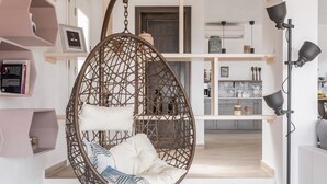 A room with a hanging chair, shelves with books, and a kitchen area in the background. - Villa Savvina (Skopelos)