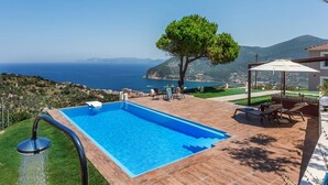 A rectangular swimming pool with a clear blue water feature, surrounded by a wooden deck and outdoor furniture. - Villa Savvina (Skopelos)