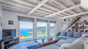 A modern living room with a large TV, a sofa, a coffee table, and a staircase leading to a balcony with a scenic view. - Villa Savvina (Skopelos)