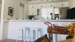 Eine moderne Küche mit weißen Schränken, einem Edelstahl-Kühlschrank, einer Mikrowelle und einem Spülbecken. Zwei weiße Barhocker und ein Korbstuhl sind ebenfalls vorhanden.