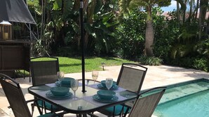 An outdoor dining area with a table and chairs, an umbrella, and a pool in the background. - Charming Floridian Home with pool located close to Lauderdale by the Sea. (Fort Lauderdale)