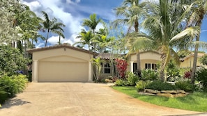 Une maison à deux étages avec un garage, entourée d’une végétation tropicale luxuriante et de palmiers.